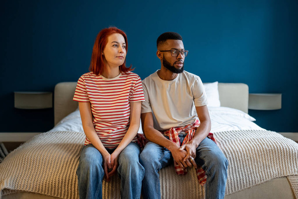 Ein breitgefächertes, multiethnisches Familienpaar sitzt auf einem bequemen Bett und träumt zusammen zum Fenster. Friedlich lächelnde Millennial-Eheleute, die sich in einer neuen Mietwohnung entspannen. Angenehm bewegungsmüde. - Foto, Bild