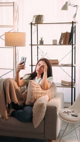 Home appointment. Video call. Flu treatment. Tired ill young girl with headache smartphone in hand in blanket on sofa with pillows in light interior with medicines on chair. - Fotoğraf, Görsel