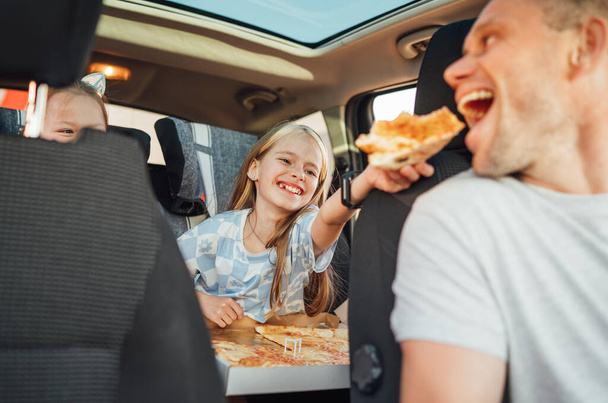 Portrait of positive smiling girl putting pizza piece in father's mouth while she sitting in child car seat . Happy childhood, fast-food eating or auto jorney lunch break concept image - Foto, immagini
