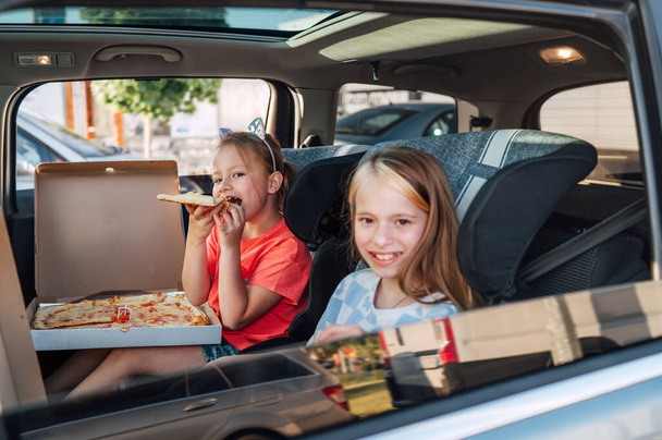 Two positive smiling sisters are happy to eat just cooked Italian pizza sitting in child car seats on the car back seat. Happy childhood, fast food eating, or auto journey lunch break concept image. - Photo, Image