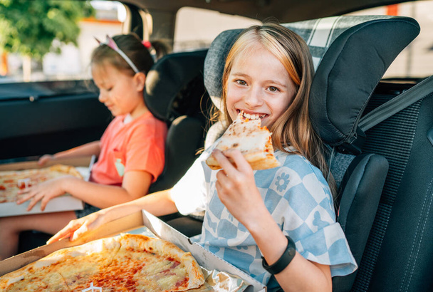 Portrait of positive smiling girl eating just cooked italian pizza sitting with a sister on car back seat in child car seats. Happy childhood, fastfood eating or auto jorney lunch break concept image. - Foto, Imagen