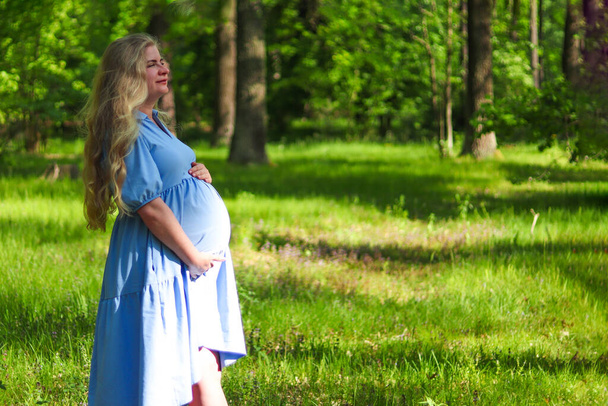 Young beautiful pregnant woman with long blonde hair holding her belly against green forest lawn. Copy space for text. Selective focus, outdoors. Concept of pregnancy and maternity. - Foto, imagen