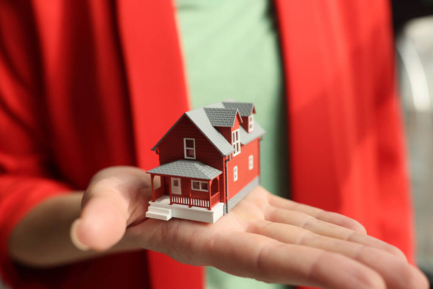 Γυναίκα μεσίτης σε σακάκι κατέχει μικρό μοντέλο σπίτι. Κόκκινο σχέδιο κτιρίου σε αντίγραφο στην παλάμη του υπαλλήλου. Έννοια της προσφοράς αγοράς ακινήτων - Φωτογραφία, εικόνα