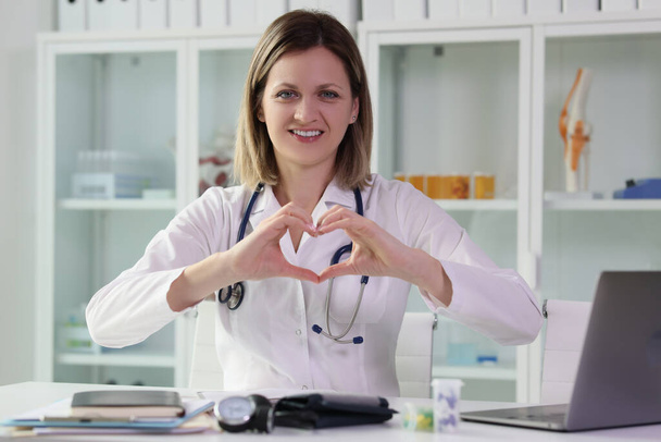 Позитивний лікар показує серце з руками, сидячи на робочому місці в клінічному кабінеті. Концепція охорони здоров'я та лікування від хвороб у медичному закладі
 - Фото, зображення