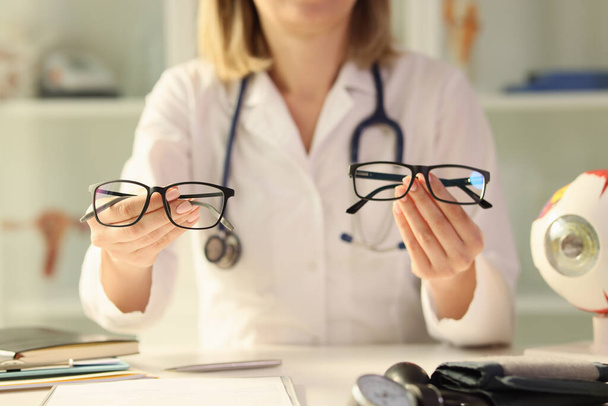 Woman optometrist compares eyeglasses models holding in hands. Female doctor offers patient options for visibility adjustment sitting at table in clinic premise - Foto, Imagem