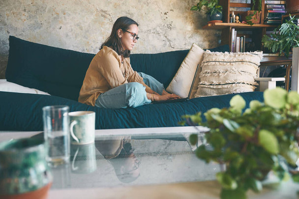 Desde el trabajo hasta el entretenimiento, la web es donde está. una mujer joven usando un portátil en el sofá en casa - Foto, Imagen