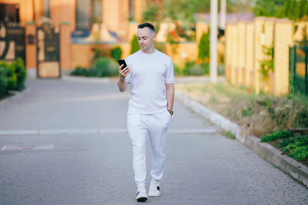 Stylish man in white costume looking at mobile phone walking at the street in the summer day - Φωτογραφία, εικόνα