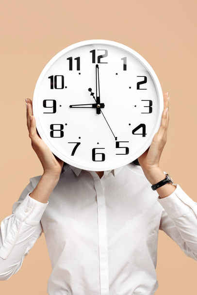 Close-up photo of young modern business woman hold a clock in front of the face over beige background - Zdjęcie, obraz