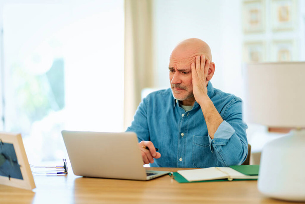Worried senior man using notebook at home. Mid aged male working online and sitting at desk. Home office. - Photo, image