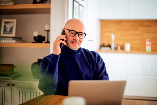 Confident middle aged man making a call and using laptop while working from home. Confident male sitting in the kitchen and having a call. Home office. - Foto, Imagen