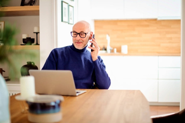 Confident middle aged man making a call and using laptop while working from home. Confident male sitting in the kitchen and having a call. Home office. - Fotó, kép