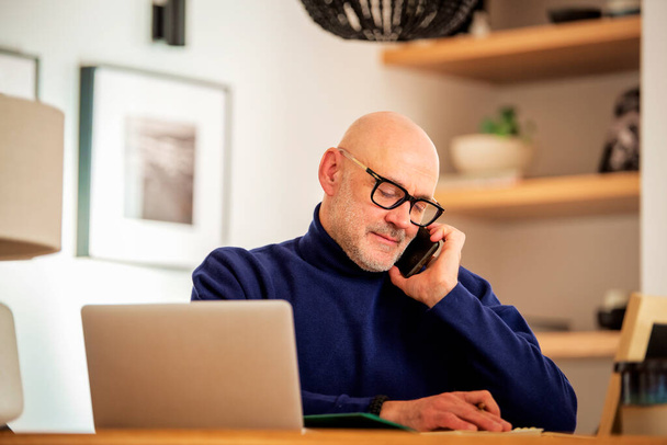 Confident middle aged man making a call and using laptop while working from home. Confident male sitting in the kitchen and having a call. Home office. - Fotoğraf, Görsel