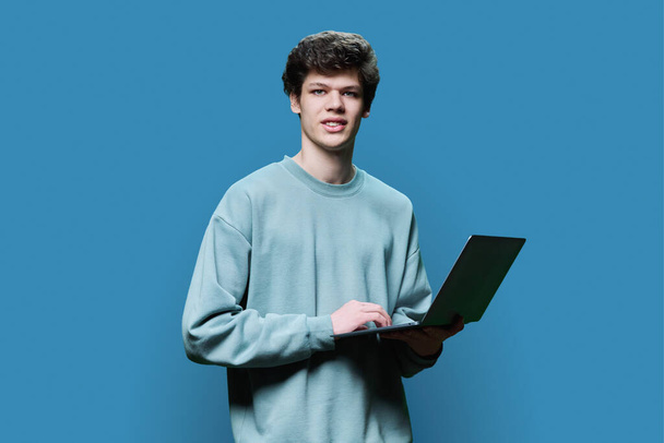 Young male college student using laptop, blue studio background. Smiling handsome guy looking at camera. Internet technologies, online learning resting, virtual remote education leisure communication - Foto, afbeelding