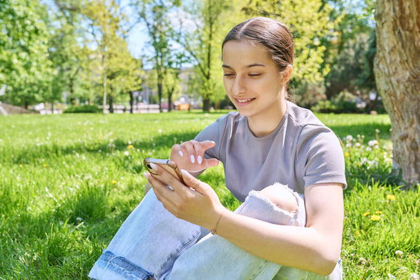 Young female sitting on the grass in the park, relaxing with smartphone. Mobile applications for leisure, learning, shopping, relaxation, online internet technology - Фото, изображение