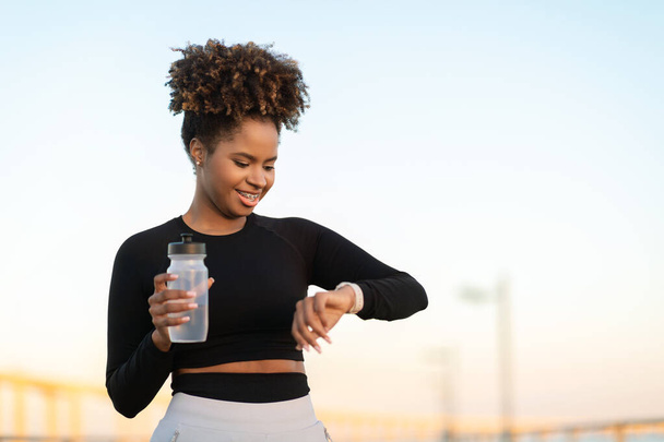 Sorridente jogger nero femminile che controlla il tempo di esecuzione su Smartwatch dopo l'allenamento all'aperto, felice giovane donna afroamericana che beve acqua e controlla i dati del fitness Tracker di guardia, copia spazio - Foto, immagini