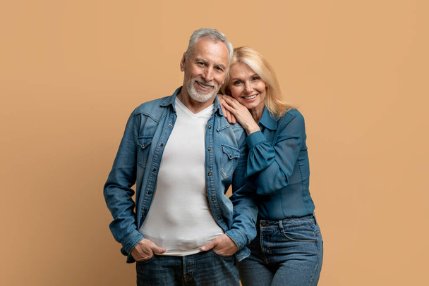 Beautiful happy elderly husband and wife wearing casual denim outfits posing together on colorful pastel background, senior couple cuddling and smiling at camera, studio shot - Photo, Image
