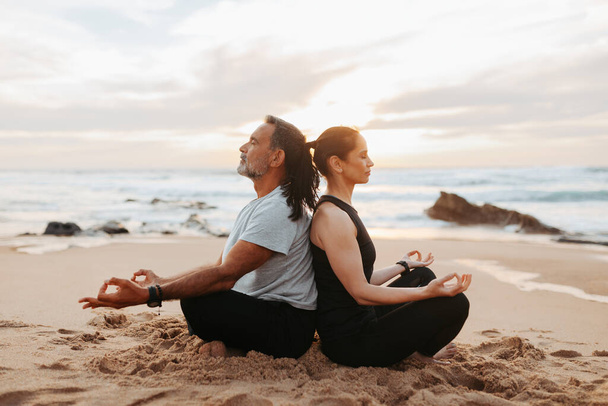 Serious calm mature european lady and man in sportswear practice yoga, meditate, enjoy workout together and peace in morning, sit on sea beach, outdoor. Rest, sports, fitness and body care - Φωτογραφία, εικόνα