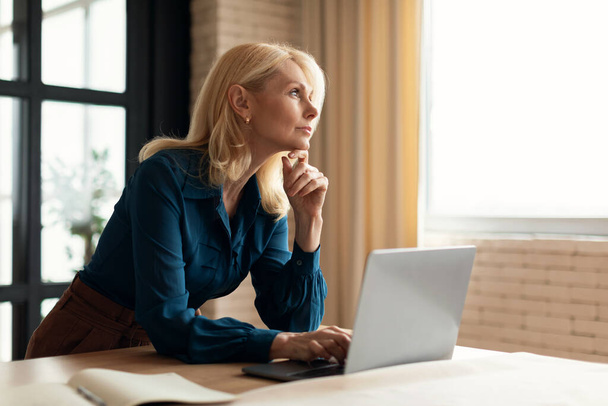 Pensive mature woman entrepreneur pondering business ideas while working at table at home, thoughtful businesswoman at workplace, leaning chin on hand and looking away - Foto, Imagem
