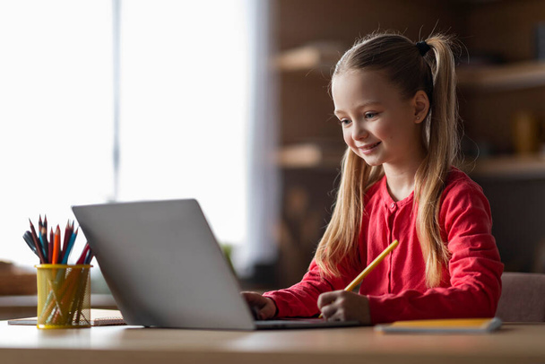Aranyos kis kaukázusi lány tanulmány laptoppal otthon és írás jegyzettömbben, mosolygó nő gyermek ül asztalnál a konyhában, tanulás a számítógéppel, élvezi az online oktatás - Fotó, kép
