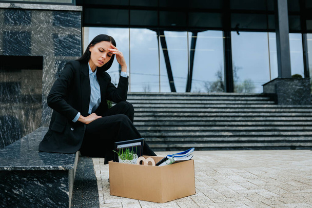 A woman sits on a cardboard box, looking upset and distressed after experiencing work loss and unemployment. Feeling lost and upset after being fired, she sits with her cardboard box. - Foto, Imagen