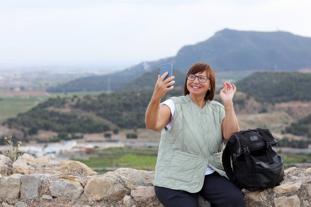 Ενηλίκων ταξιδιωτική blogger γυναίκα με σακίδιο στέκεται στο λόφο μετά την πεζοπορία μέχρι κατέχει smartphone, επικοινωνεί με τους οπαδούς της και δείχνει βουνά στα μέσα κοινωνικής δικτύωσης. Ασφάλεια σόλο γυναικείο σακίδιο στην Ευρώπη. - Φωτογραφία, εικόνα