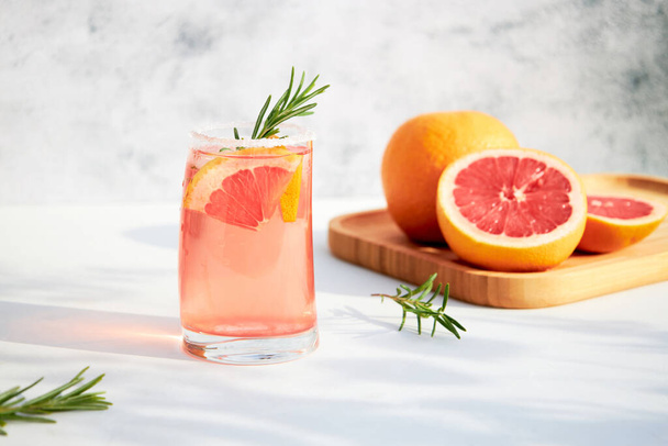 Elegáns üveg friss, hűsítő grépfrútból vagy vörös narancsból készült koktéllal, jégkockákkal és egy csipetnyi rozmaringgal szürke alapon egy fényes, napos napon. Nyaralás és nyári italok koncepciója - Fotó, kép