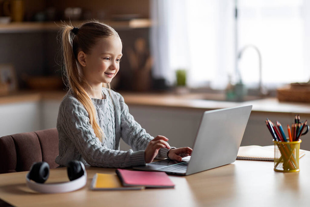 Κωδικοποίηση για παιδιά. Χαριτωμένο κοριτσάκι χρησιμοποιώντας το φορητό υπολογιστή, ενώ κάθεται στο γραφείο στο σπίτι, χαμογελώντας Preteen θηλυκό παιδί παρακολουθούν μαθήματα προγραμματισμού, κοιτάζοντας οθόνη υπολογιστή και πληκτρολογώντας στο πληκτρολόγιο - Φωτογραφία, εικόνα
