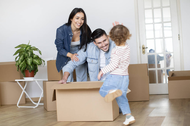 Cheerful Family Of Three Having Fun While Relocating Home, Happy Young Parents And Their Toddler Son Fooling Together While Unpacking Things On Moving Day, Playing With Cardboard Boxes In Living Room - Photo, Image