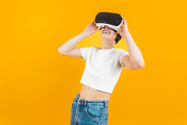 Keskikokoinen kuva nuoresta hymyilevästä naisesta valkoisessa topissa ja farkuissa leikkimässä VR-kuulokkeissa. Keltainen tausta. Laadukas kuva - Valokuva, kuva