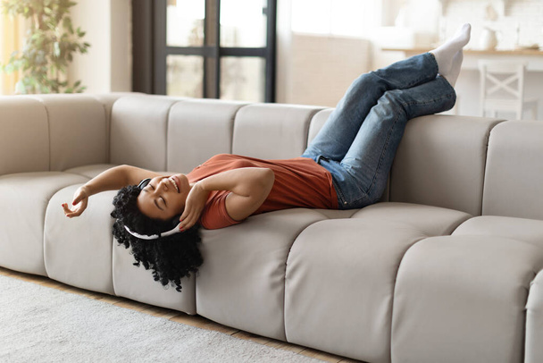 好きなパスタイム。自宅で楽しいアフリカ系アメリカ人女性が自宅で、リビングルームで休憩し、無料のスペースで音楽を聞きながらソファに横たわっている陽気な若い黒人女性 - 写真・画像