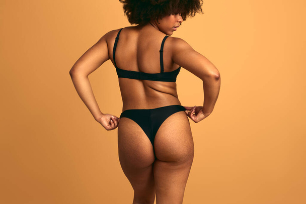Back view of African female plus size model touching panties while standing against orange background in studio - Foto, immagini