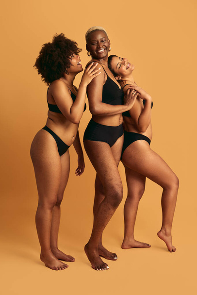 Full body of happy young multiracial female models in black underwear standing against yellow background and smiling while touching each other in studio - Φωτογραφία, εικόνα