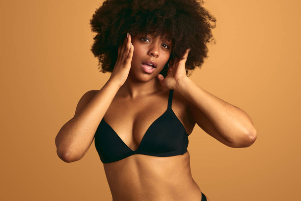 Young African female in black bra with Afro hair and bare belly touching cheeks while opening mouth against orange background - Foto, Bild