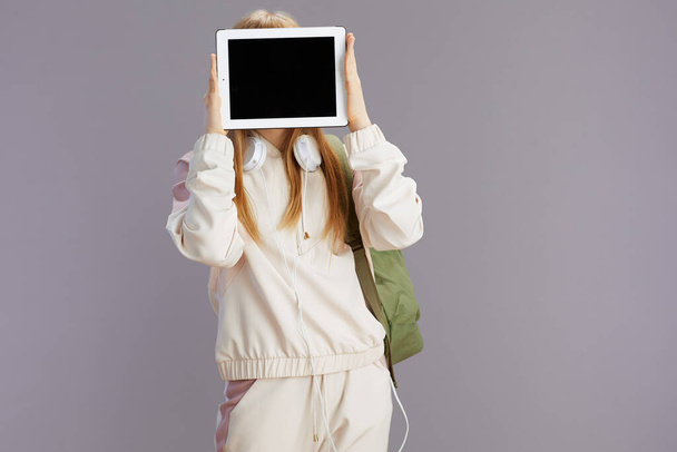 modern lány bézs tréningruha hátizsák és fejhallgató mutatja tabletta PC üres képernyő elszigetelt szürke háttér. - Fotó, kép