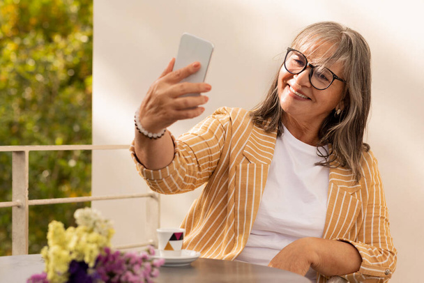 Cheerful old caucasian gray-haired woman in glasses sits at table in cafe, make selfie, has video call on phone, outdoor. Blog, app, rest, communication remotely and social networks - Photo, Image