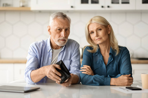 Financial Problems. Upset Senior Couple Showing Empty Wallet While Sitting At Table In Kitchen, Mature Husband And Wife Suffering Retirement Crisis, Having Economy Isuues, Looking At Camera - Photo, Image