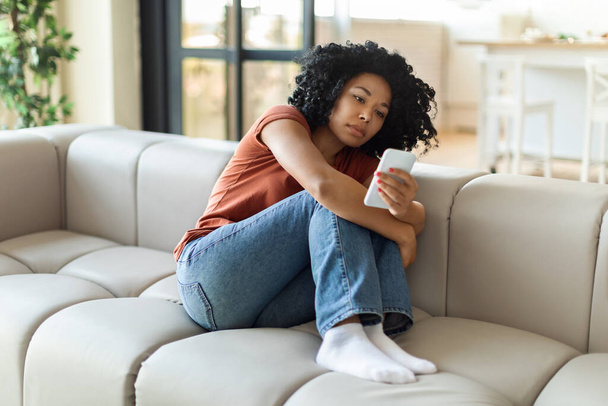Upset black woman looking at cellphone screen while sitting on couch at home, depressed african american lady received bad news, reading unpleasant message or waiting for important call, copy space - Foto, imagen