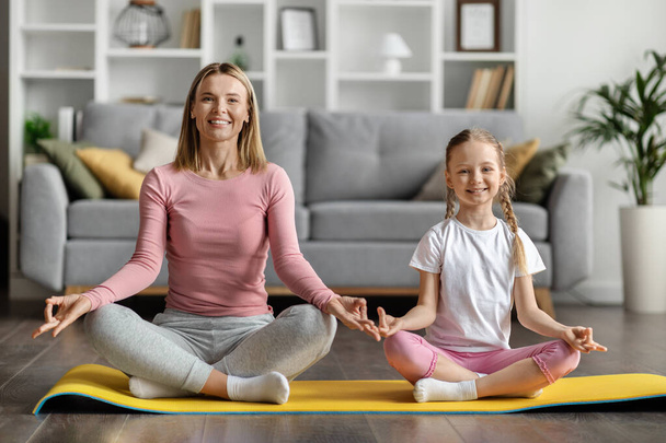 Cute Little Girl And Her Young Mother Meditating Together At Home, Smiling Caucasian Mom And Adorable Female Child Sitting In Lotus Position, Training On Fitness Mat In Living Room, Smiling At Camera - Foto, immagini
