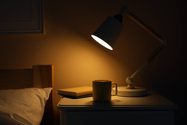 Stylish modern desk lamp, books and cup of drink on white nightstand in dark bedroom - 写真・画像