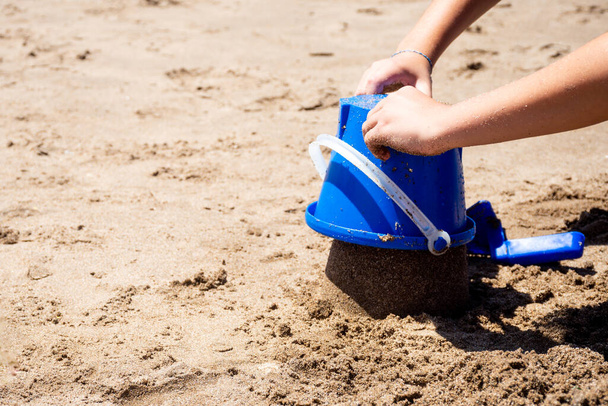 Hands of a child playing with a shovel and a blue plastic bucket in the sand on the beach. - Foto, Imagen