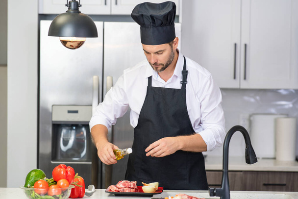 Handsome man cooking in kitchen. Mature man cooking with vegetables and meat in kitchen interior. Caucasian man in apron and chef hat cook tasty lunch in kitchen. Healthy food for dinner - 写真・画像