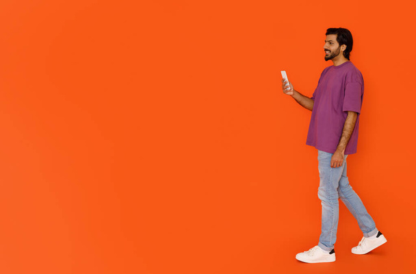 Coole, gut aussehende junge Ostler in stylischem Outfit, die mit Smartphone spazieren gehen und auf orangefarbenem Studiohintergrund lächeln, mit netter mobiler App, Panorama mit Kopierplatz für Werbung - Foto, Bild