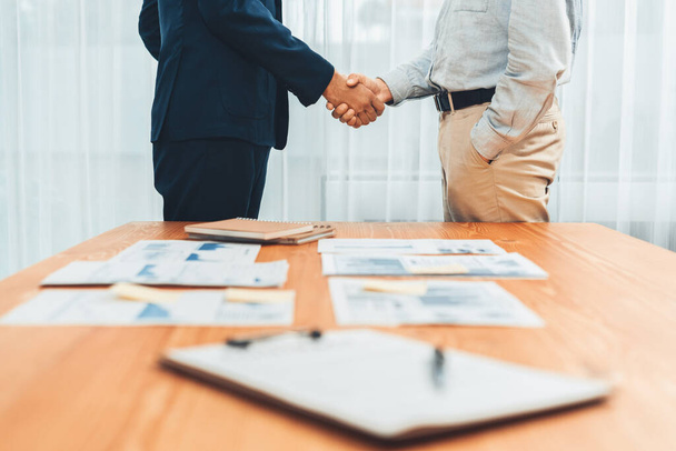 Two businesspeople shake hand after signing contract document to merge their partnerships in conference room and finalized pile of papers of financial report and data analysis on meeting table. Entity - 写真・画像