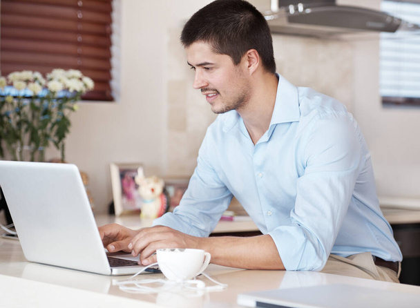 Genießen Sie die Vorteile des Internets. Ein gutaussehender Mann sitzt in seiner Küche, mit einer Tasse Kaffee auf seinem Laptop - Foto, Bild