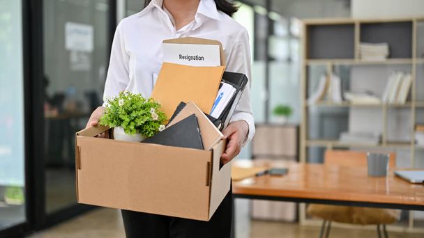 Обрезанное изображение работницы офиса стоит в офисе с картонной коробкой со своими вещами и заявлением об отставке. уйти в отставку, оставить работу, уволен, безработный - Фото, изображение