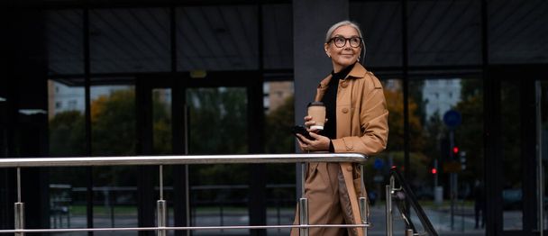 foto orizzontale di un capo donna matura di successo con una tazza di caffè tra le mani sullo sfondo dell'edificio. - Foto, immagini