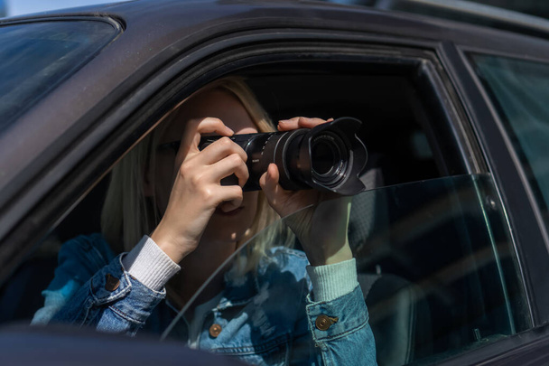 Young blonde woman in sunglasses takes pictures on a professional camera while sitting in a car. Concept of journalism, detective, papparation - Fotografie, Obrázek