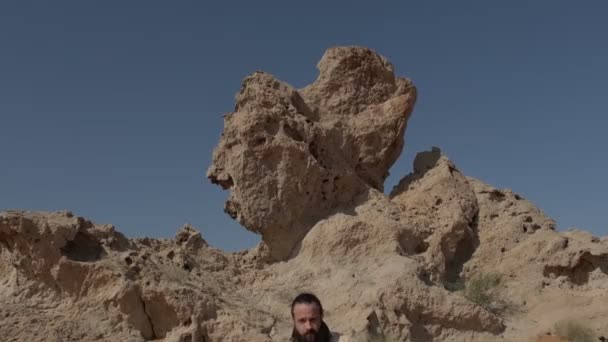 Um jovem xamã barbudo está na areia entre dunas e rochas contra um céu limpo. Movimento lento - Filmagem, Vídeo