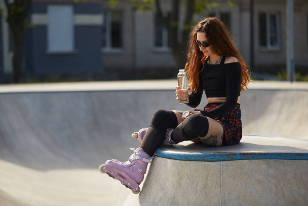 Roller blader female sitting on a ramp top in a skate park with a glass bottle of water in hand. Cool young skater wearing modern aggressive inline skates on feet - Фото, изображение