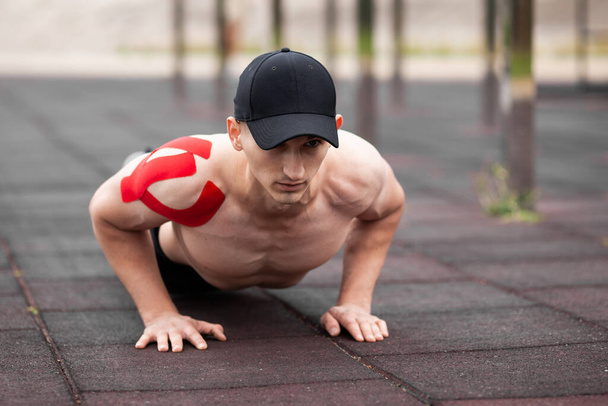 Young European athlete with injury is training outdoors. Kinesiology ( therapeutic, Kinesio, k-tape, KT, physio) tape on shoulder. Rehabilitation and recovery. Push up from the floor. - Photo, image